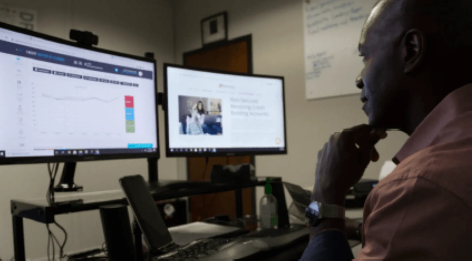A man in a pink shirt studies graphs on dual monitors in an office.