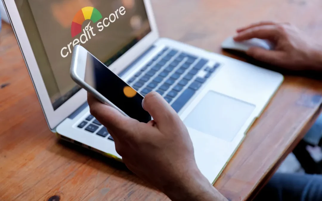 A person checks their credit score on a laptop while holding a smartphone, seated at a wooden table.