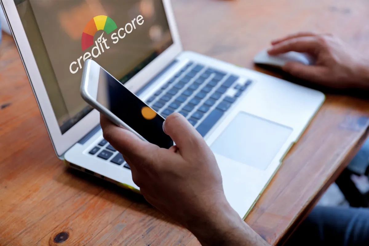 A person checks their credit score on a laptop while holding a smartphone, seated at a wooden table.