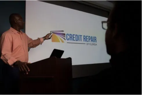 A presenter points to a "CREDIT REPAIR OF FLORIDA" slide in a dimly lit presentation room.