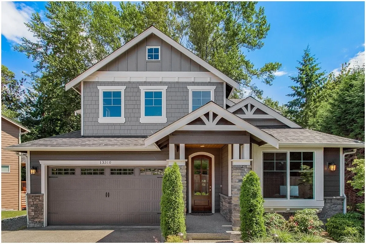 A two-story house with a garage, stone accents, and greenery under a blue sky.
