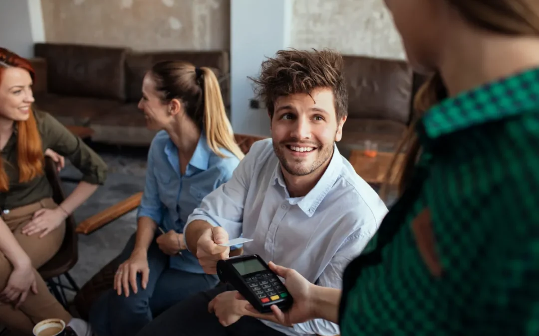 Cheerful man hands over a credit card, with women chatting in the background, in a social setting.