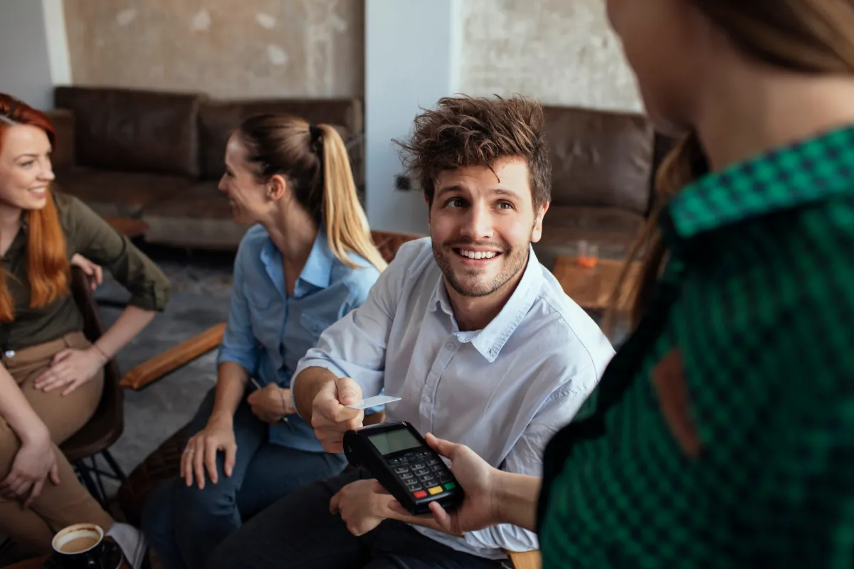 Cheerful man hands over a credit card, with women chatting in the background, in a social setting.