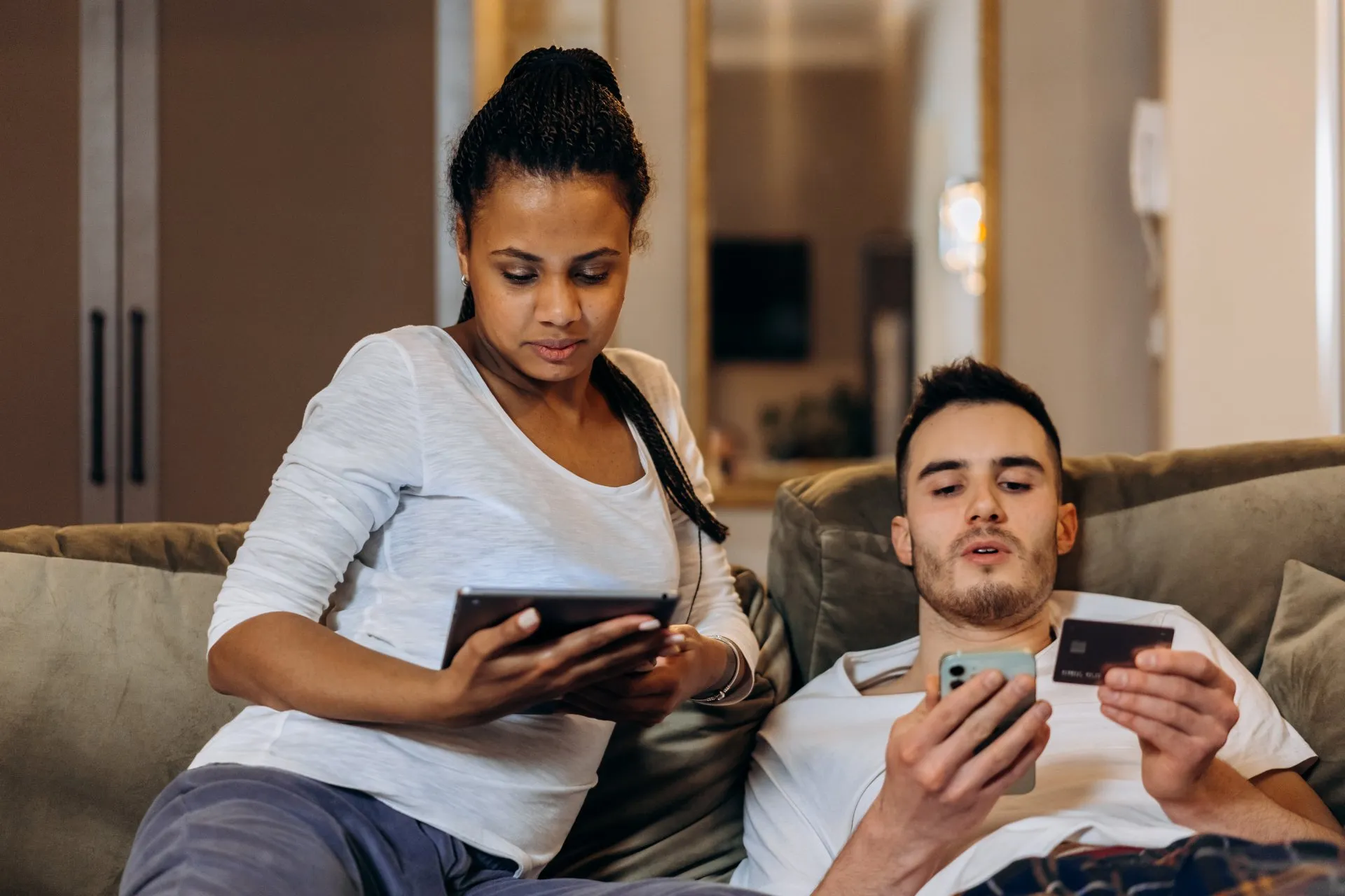 Couple on a couch using a tablet and a smartphone with a credit card.