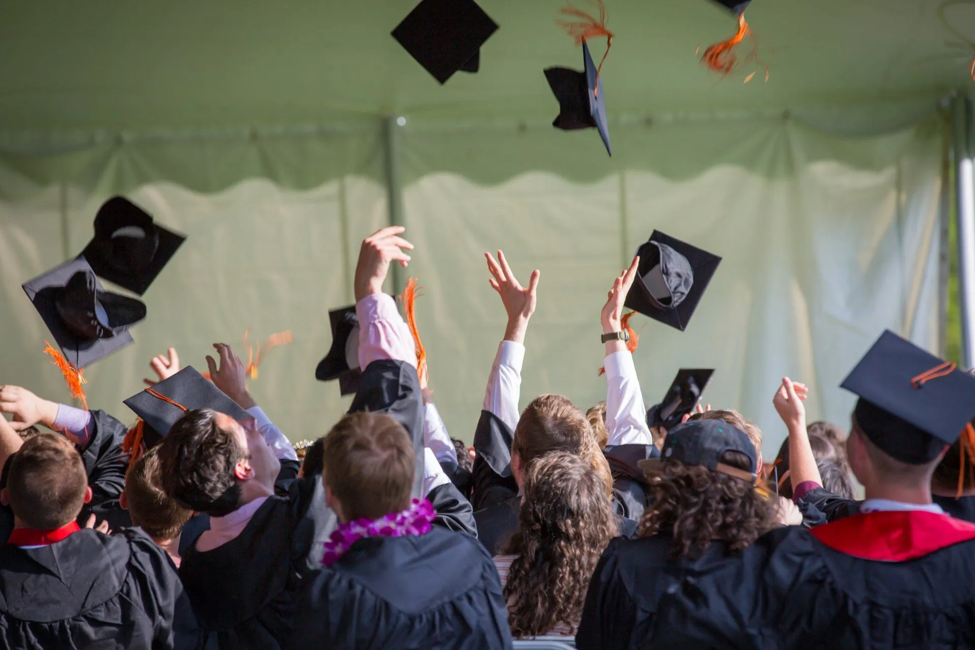 Graduates tossing their caps in celebration, with a tent blurred in the background.