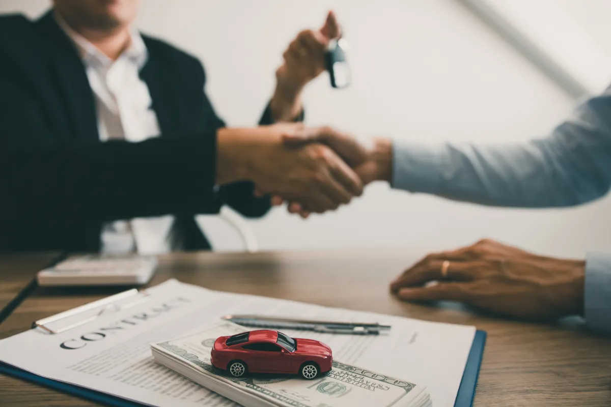 Handshake over a table with a contract, toy car, and cash, suggesting a vehicle transaction.