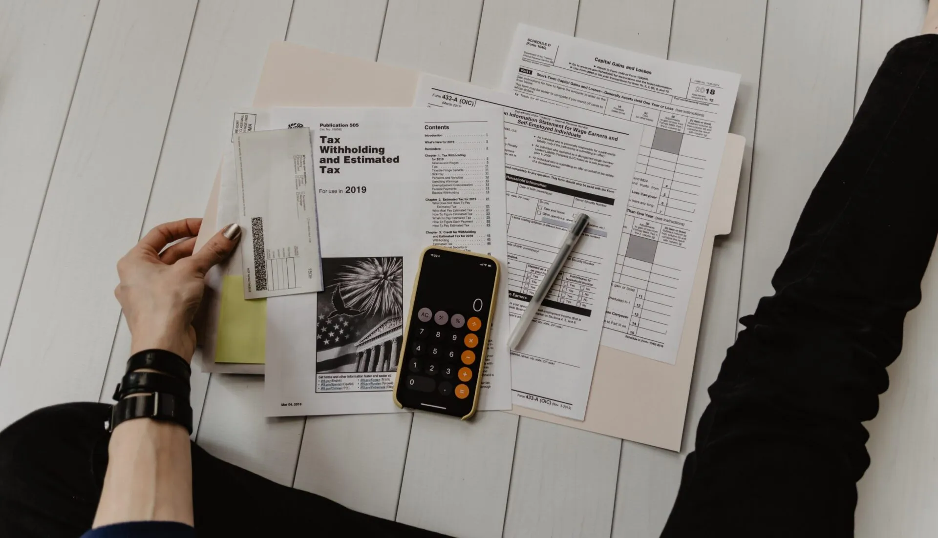 Overhead view of someone organizing tax forms with a calculator and pen on a white wooden surface.