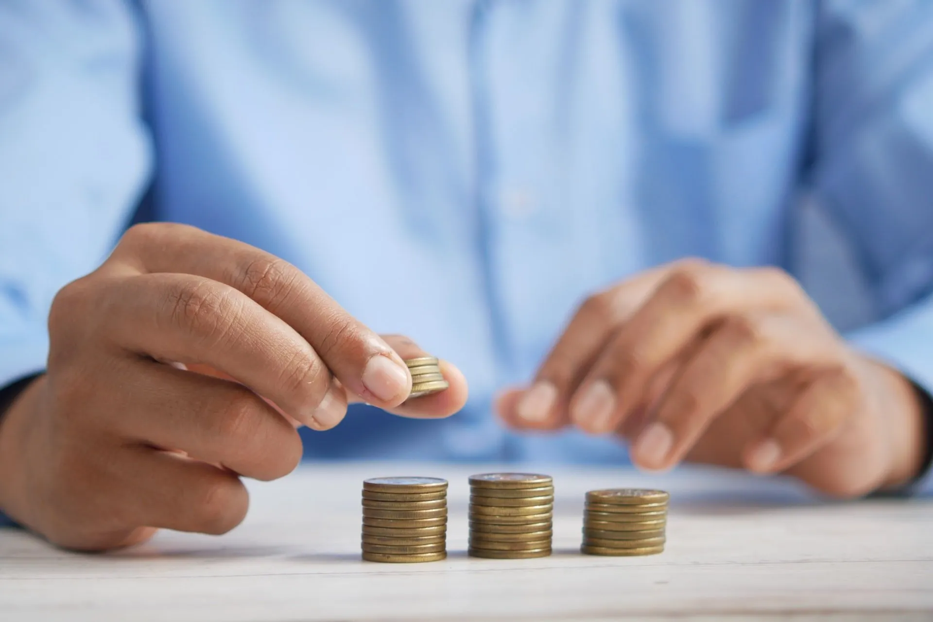 Person in a blue shirt is stacking coins in piles on a table, symbolizing money management or savings.