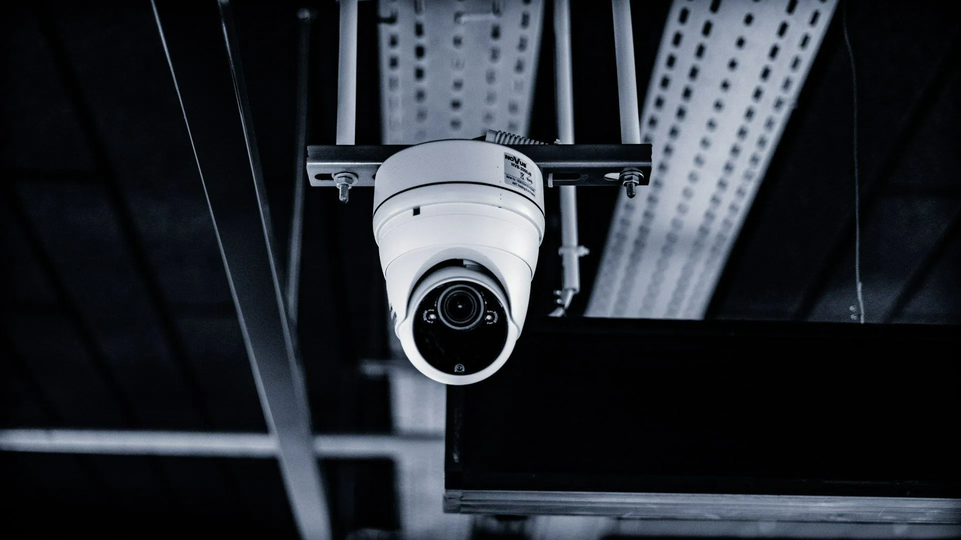 Security camera mounted on a ceiling, focusing downwards, with industrial elements in the background.