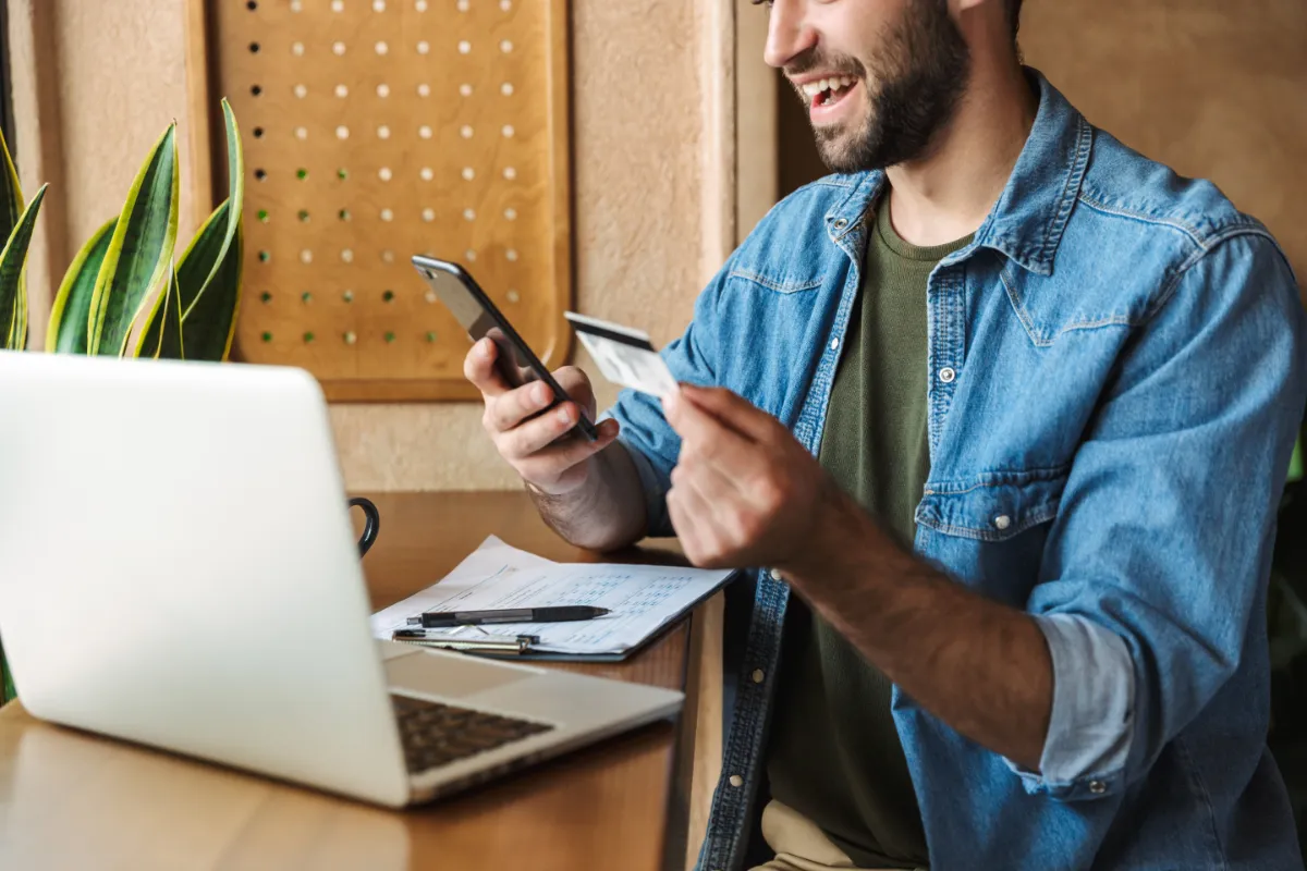 Smiling man using a smartphone and holding a credit card, with a laptop and papers on the desk