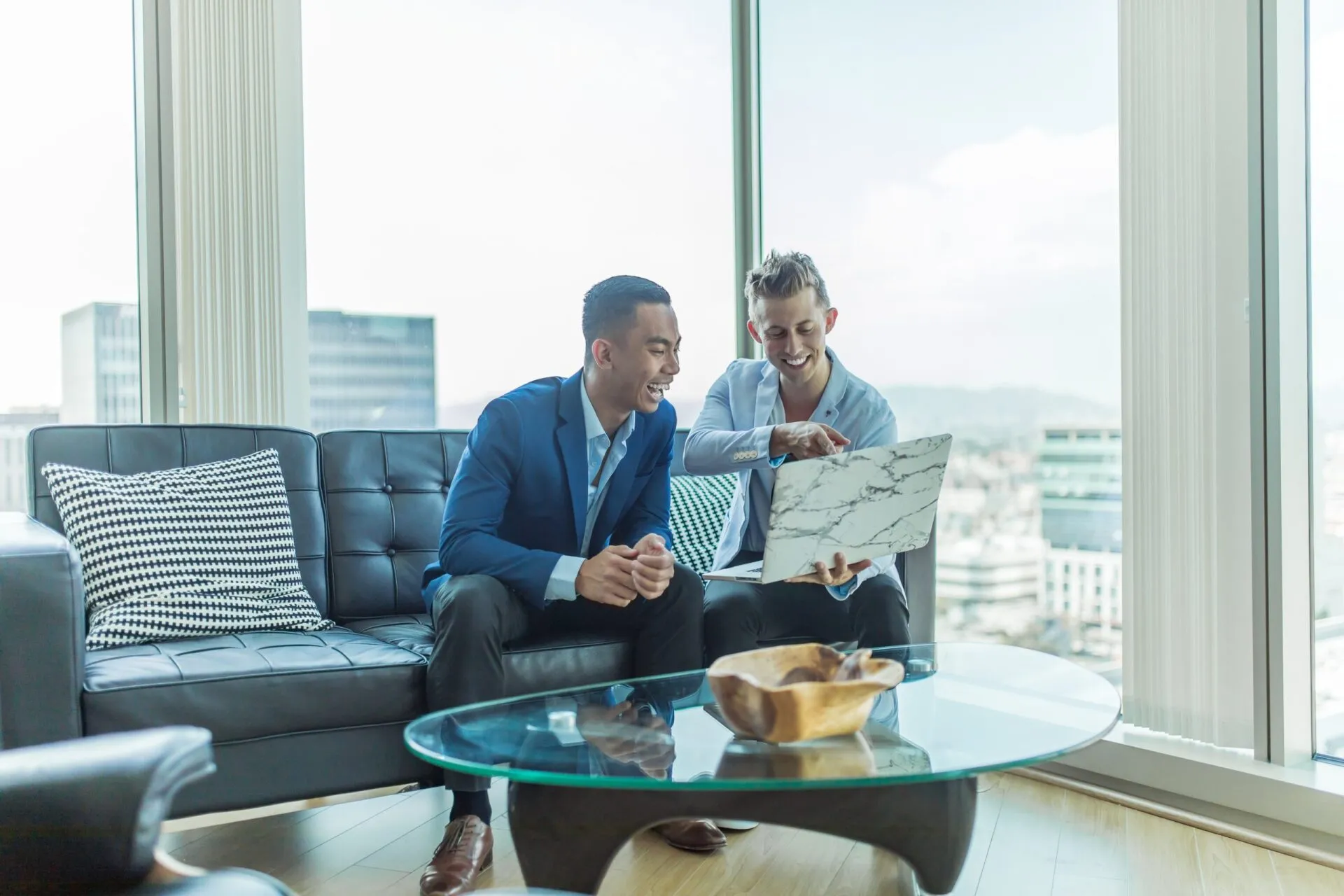 Two smiling men reviewing a laptop in a well-lit room with cityscape views.