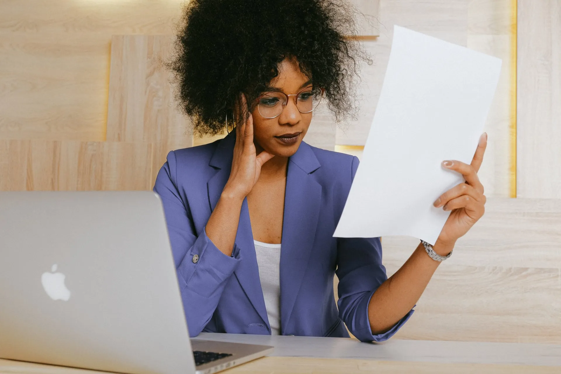 woman in a blue blazer reviews a document at her desk with a laptop open beside her.