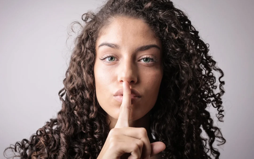 Woman with curly hair putting her finger to her lips in a gesture for silence.