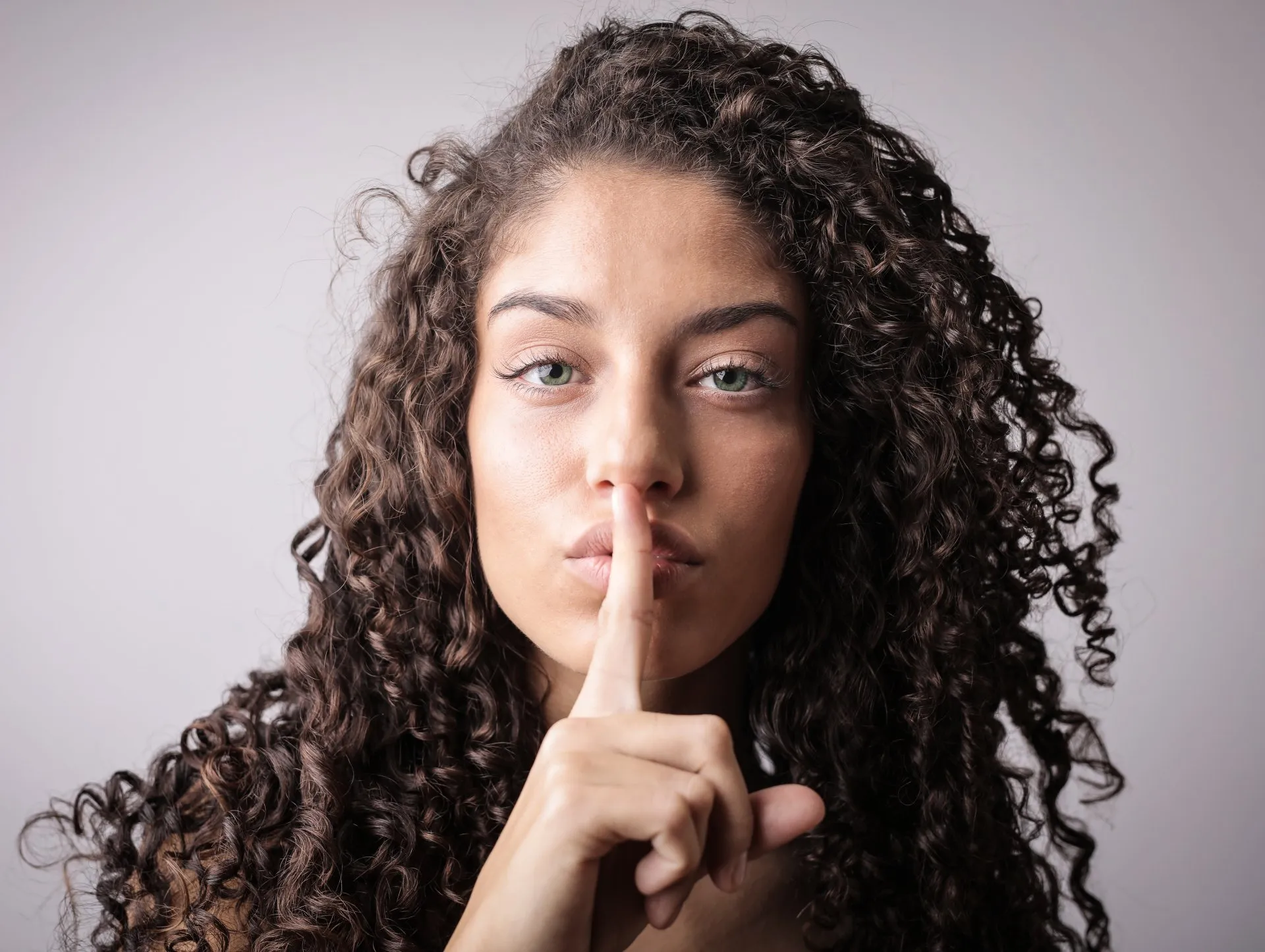 Woman with curly hair putting her finger to her lips in a gesture for silence.